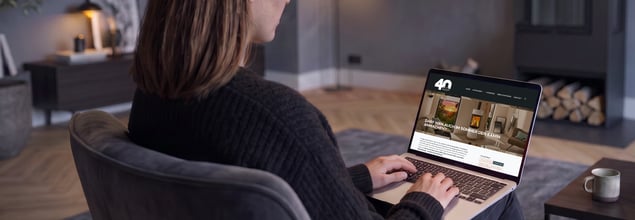 Eine Frau sitzt mit einem Laptop auf dem Schoß, auf dem der Blogbeitrag "Darf man im Sommer den Kamin anmachen?" geöffnet ist, in einem Sessel im Wohnzimmer.