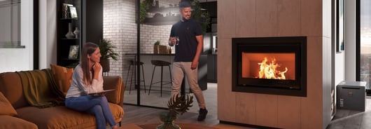 Eine Frau sitzt im Wohnzimmer auf einer Couch, ein Mann steht daneben, sie unterhalten sich und blicken beide auf den Kaminofen.