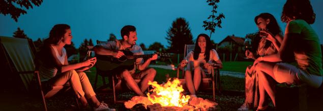Fünf Personen sitzen in der Dämmerung um ein Lagerfeuer, trinken Wein, singen und eine Person spielt Gitarre.