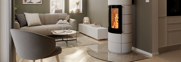 Ein weißer Kaminofen mit Glasvorlegeplatte steht in einem hell eingerichteten Wohnzimmer neben einer Couch mit Tisch und einem Stuhl.