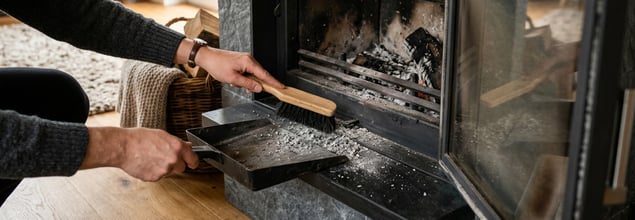 Jemand reinigt den Feuerraum eines Kaminofens mit einem Kehrblech.