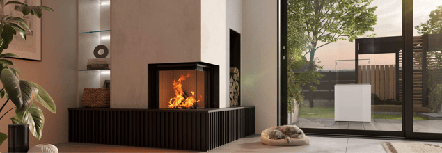 Ein weißer Kaminofen mit Holzfach steht in der Ecke eines Wohnraumes direkt neben einer großen Fensterscheibe mit Blick in den Garten.