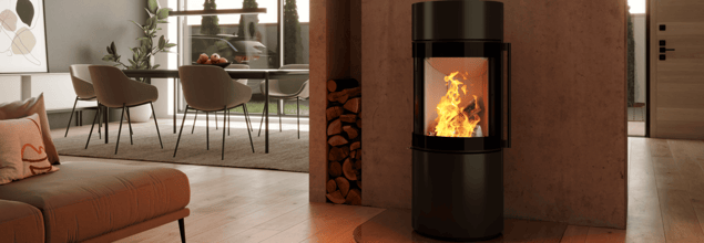 Ein schwarzer Kaminofen mit brennendem Feuer steht vor einer Betonwand mit Holzfach, welche als Raumteiler von Wohn- und Essbereich dient.