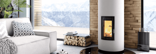 Ein weißer Kaminofen mit einem Holzfach daneben steht vor einer gemauerten Wand zwischen zwei großen Fenstern in einem modernen Wohnzimmer.