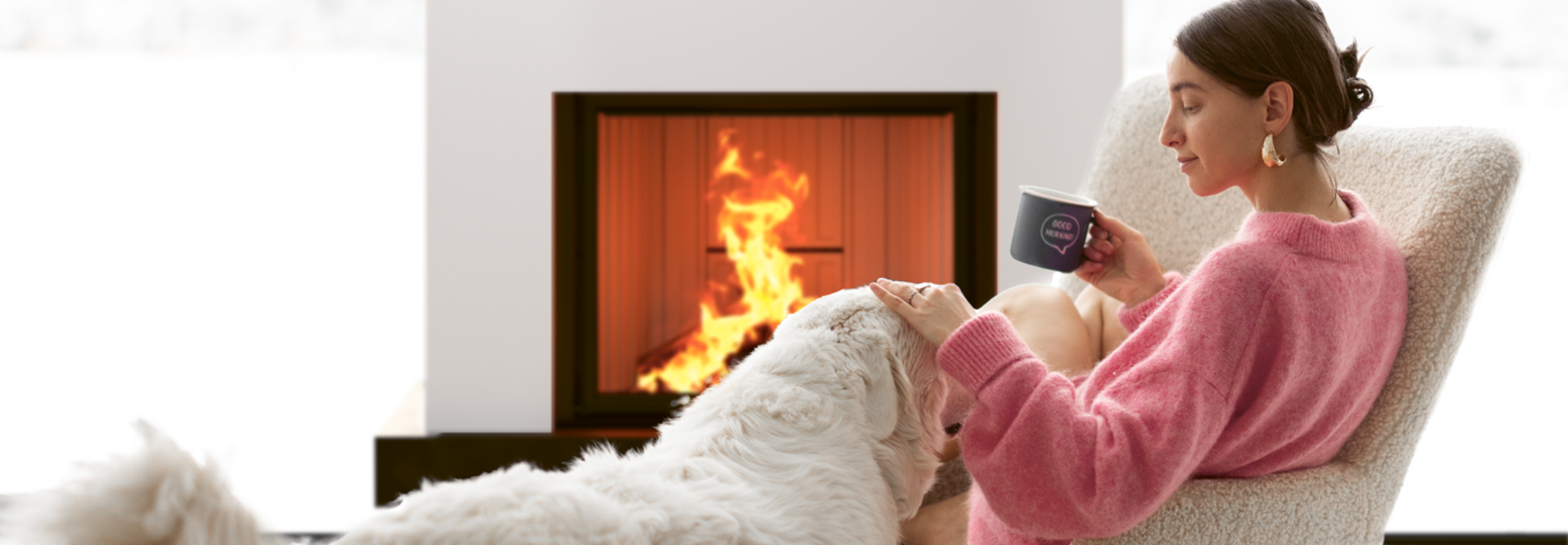 Eine Frau sitzt mit einer Tasse in der Hand auf einem Sessel vor einem Kaminofen und streichelt einen Hund.