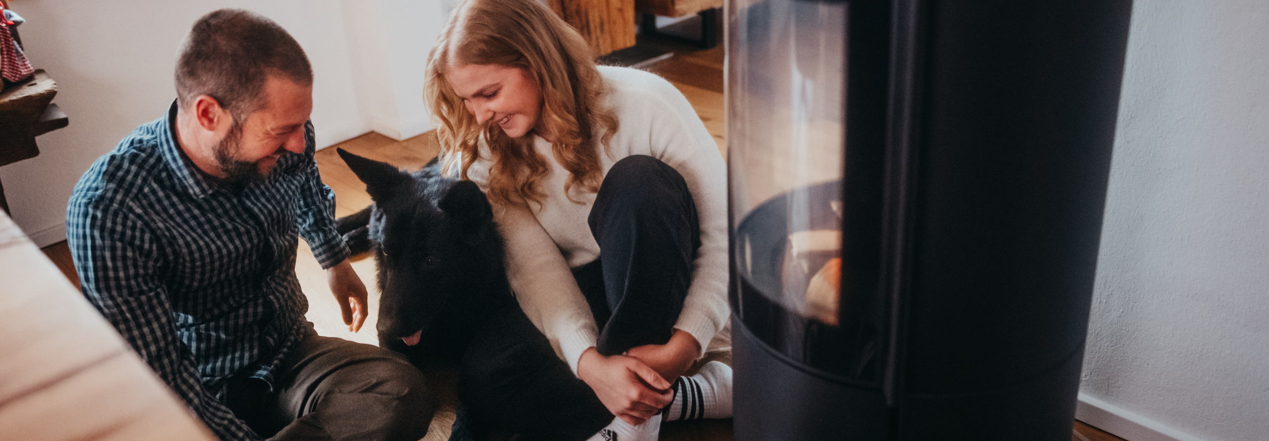 Paar sitzt mit schwarzem Hund vor einem Kamin und genießt die Wärme.
