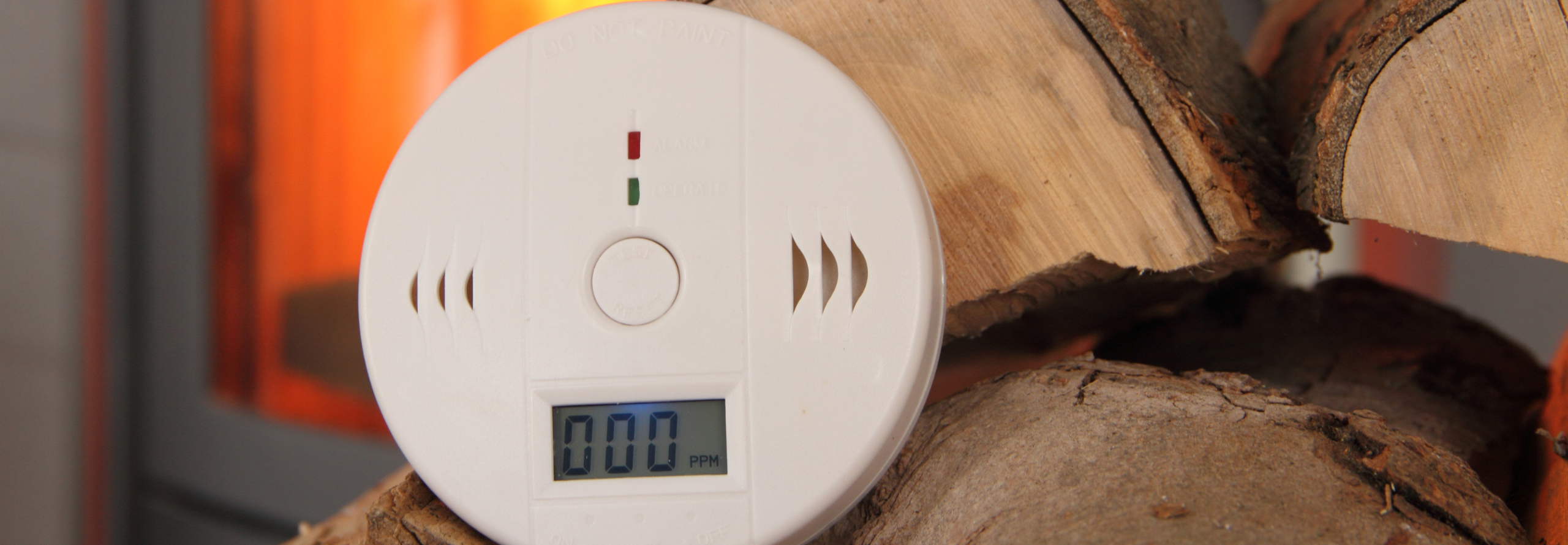 Bild eines Rauchmelders, der auf gestapelten Holzscheiten vor einem Ofen liegt.